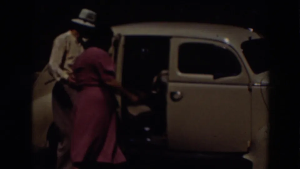 A man in a brimmed hat opens the door of a light-colored vintage car as a woman in a pink dress approaches during the 1937 outing.