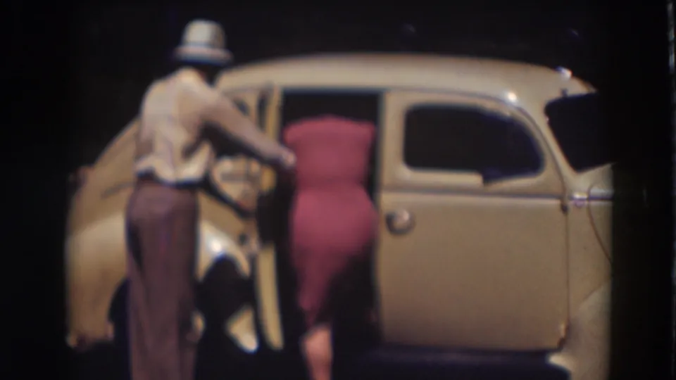 Beside the light-colored car, the man reaches toward the open door while the woman in the pink dress leans in, continuing the departure sequence.