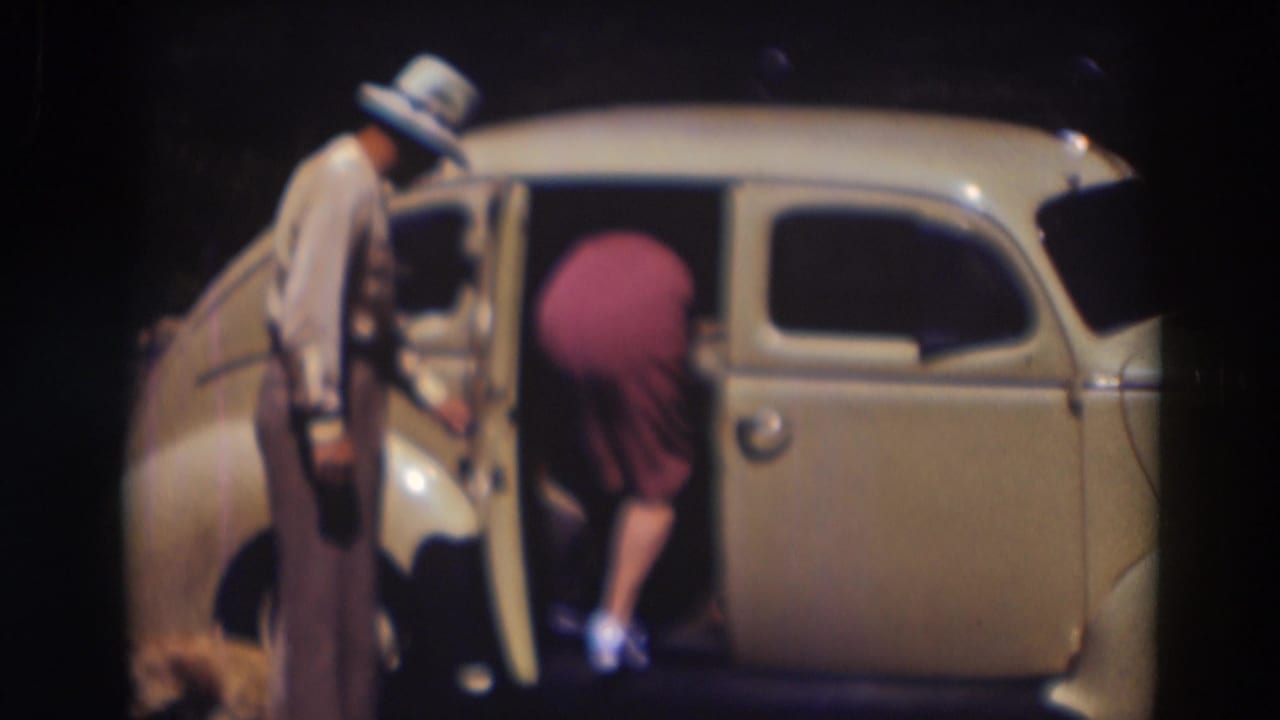Man in fedora holds an open car door as a woman in a pink dress leans into the vehicle.