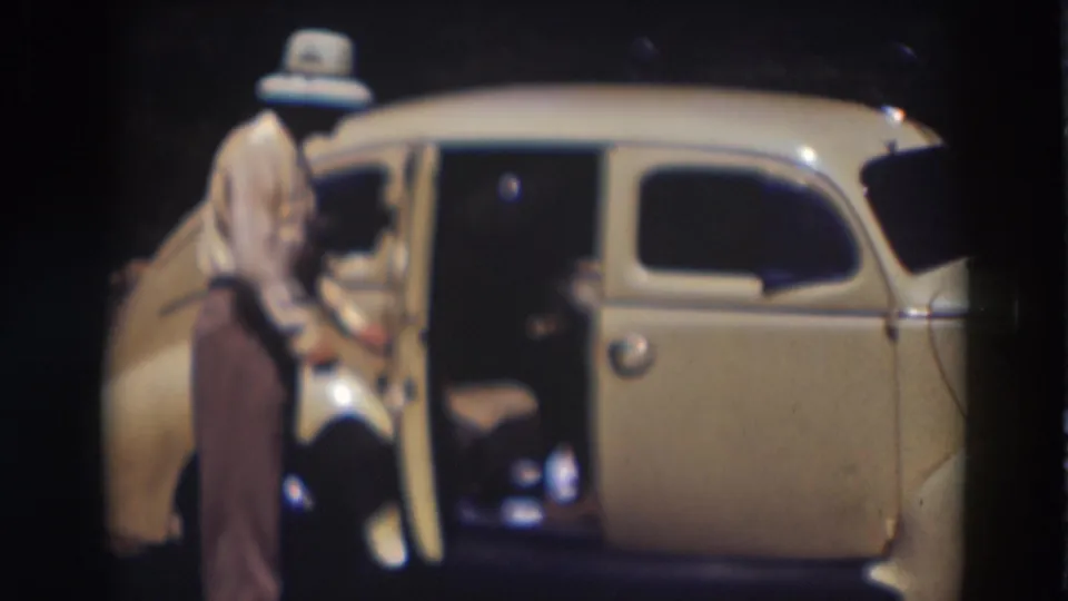 Man in fedora and brown trousers stands by an open passenger door of a cream-colored car in 1937 Truckee footage.