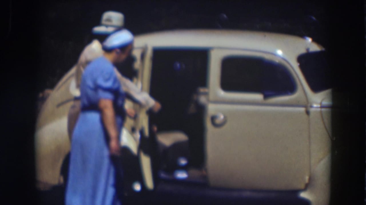 Woman in a blue dress stands beside an open rear door of a cream vintage car as a man assists nearby.