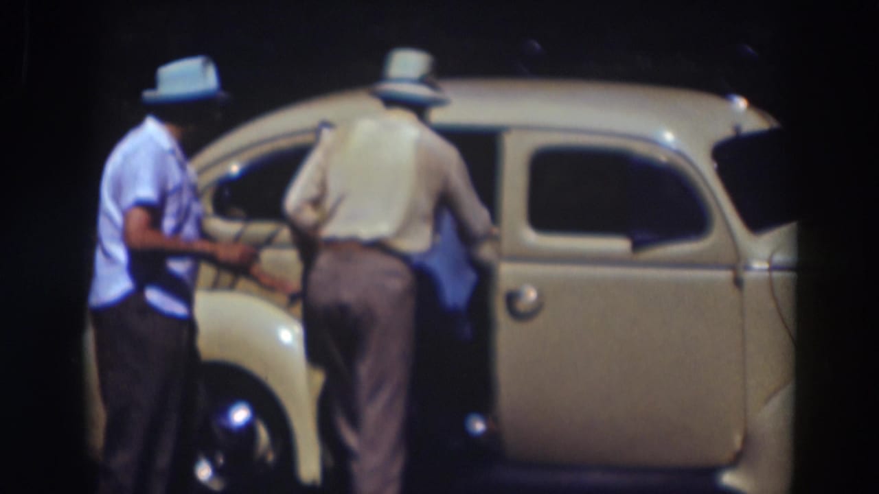 Two men in hats stand beside an open rear door of a cream-colored car at night.