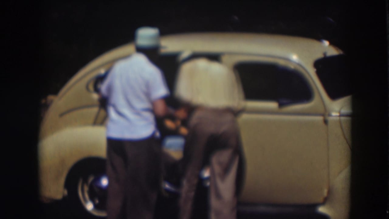 Two men face the side of a pale yellow car while one reaches into the open doorway.