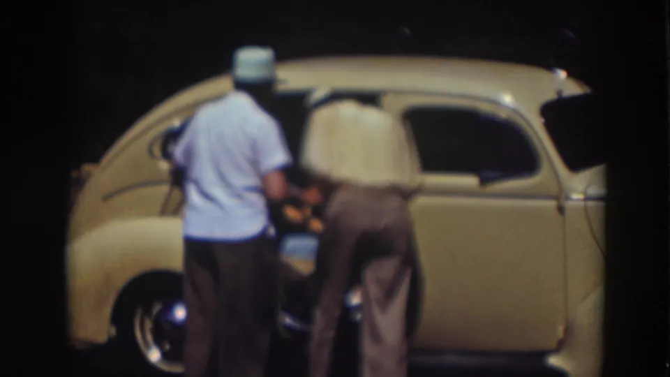 Two men in light shirts and hats stand close to a pale yellow compact car as one leans toward the open door.