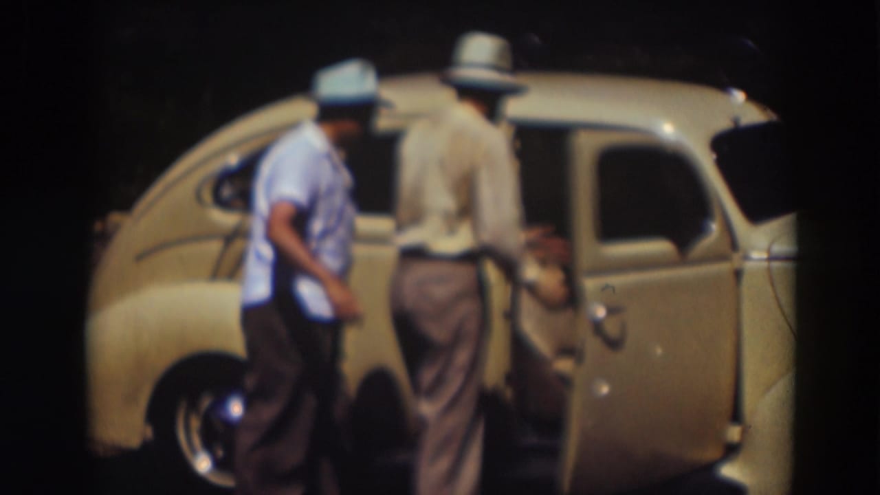 One man steps aside while another stands at the side of the pale yellow car, continuing the effort to enter it.