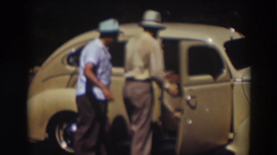One man steps aside while another stands at the side of the pale yellow car, continuing the effort to enter it.