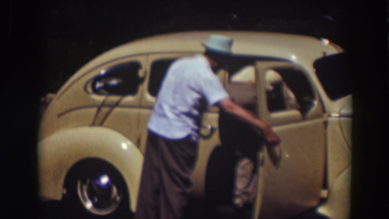 Man in a light shirt and hat closing the door of a cream-colored rounded car.