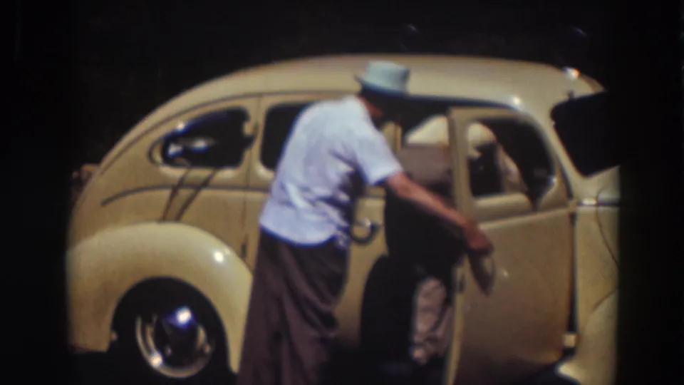 In 1937 Truckee, California, a man in a light shirt and fedora stands beside a cream-colored rounded car with the door open.