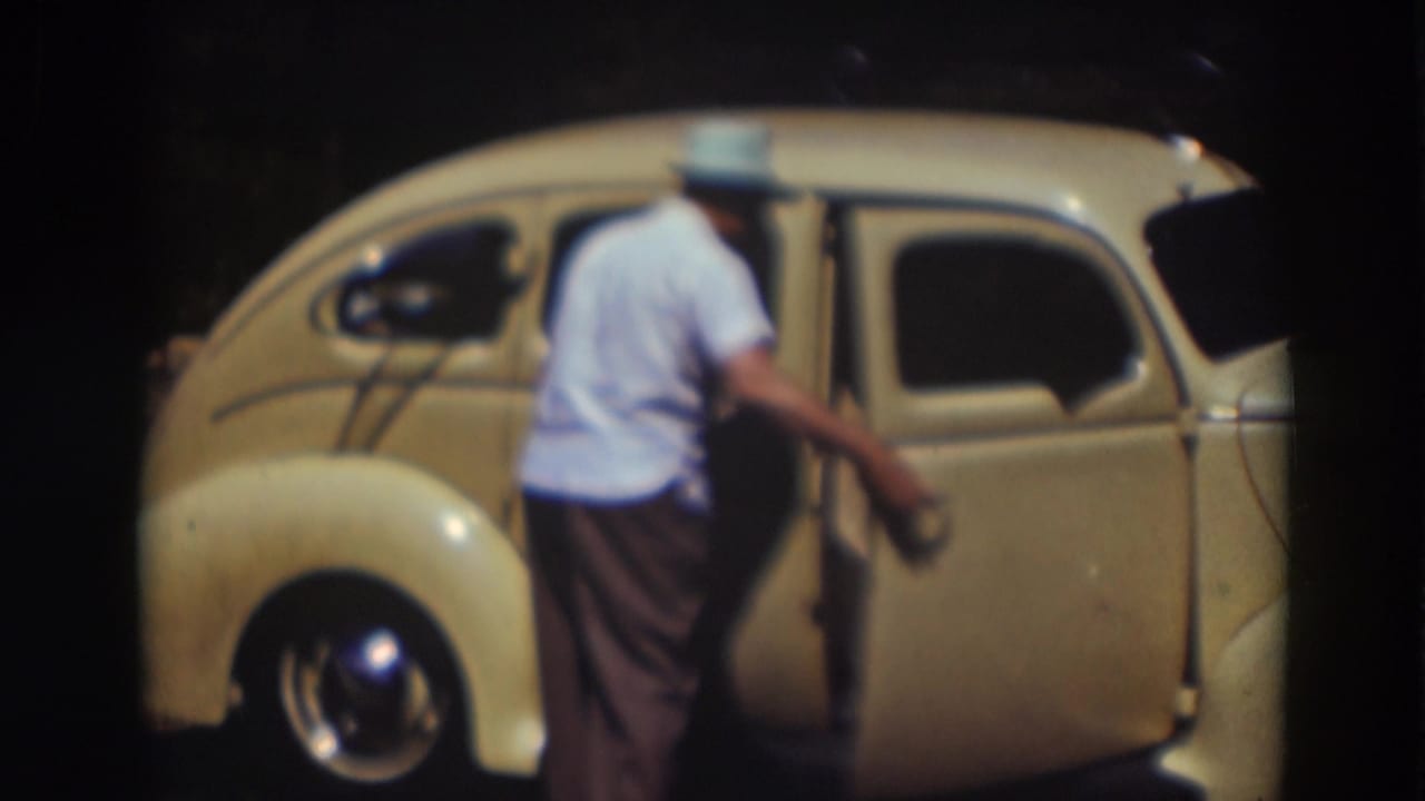 A man in a light shirt and hat stands beside the closed door of a cream-colored rounded car in the Truckee home-movie footage.