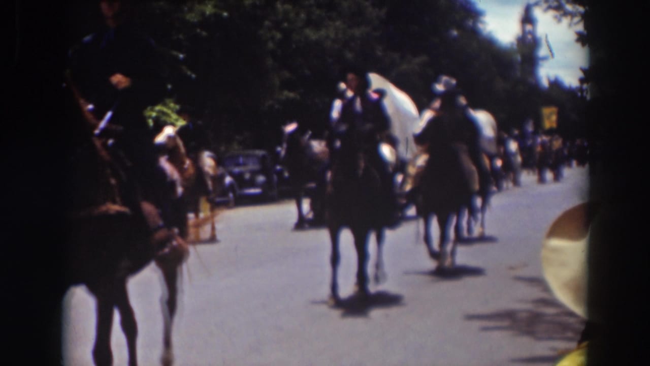 Horseback riders approach on a tree-lined road with covered wagons and cars behind them.