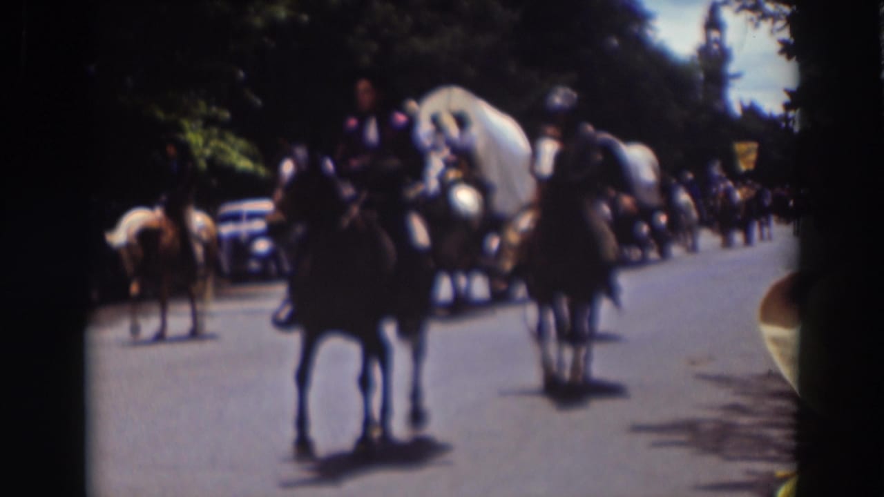 A rider in period-style attire passes close to the camera during the Truckee procession, with covered wagons and parked cars farther back.