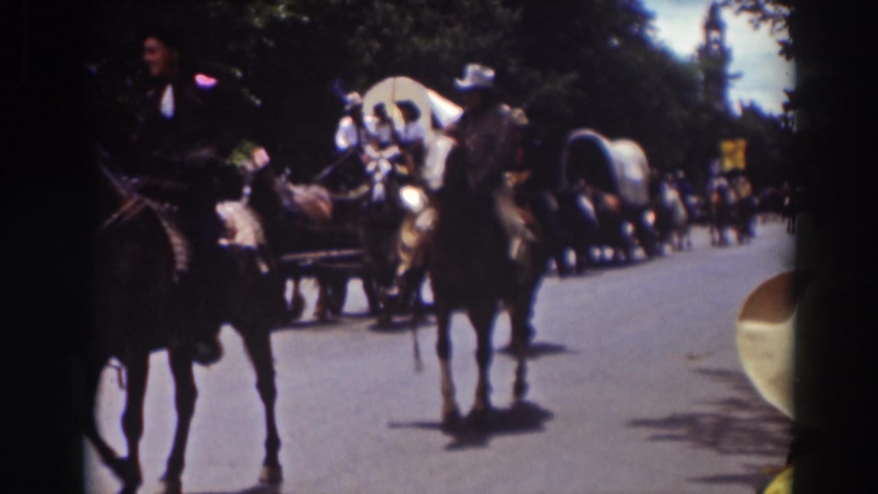 Truckee parade scene with riders on horseback moving past the camera, covered wagons behind them, and spectators along the roadside.