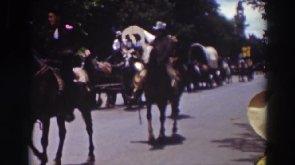 Truckee parade scene with riders on horseback moving past the camera, covered wagons behind them, and spectators along the roadside.