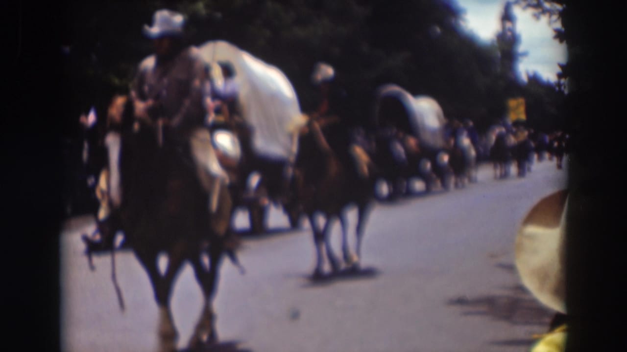 Horseback riders and covered wagons pass along a street during a parade.