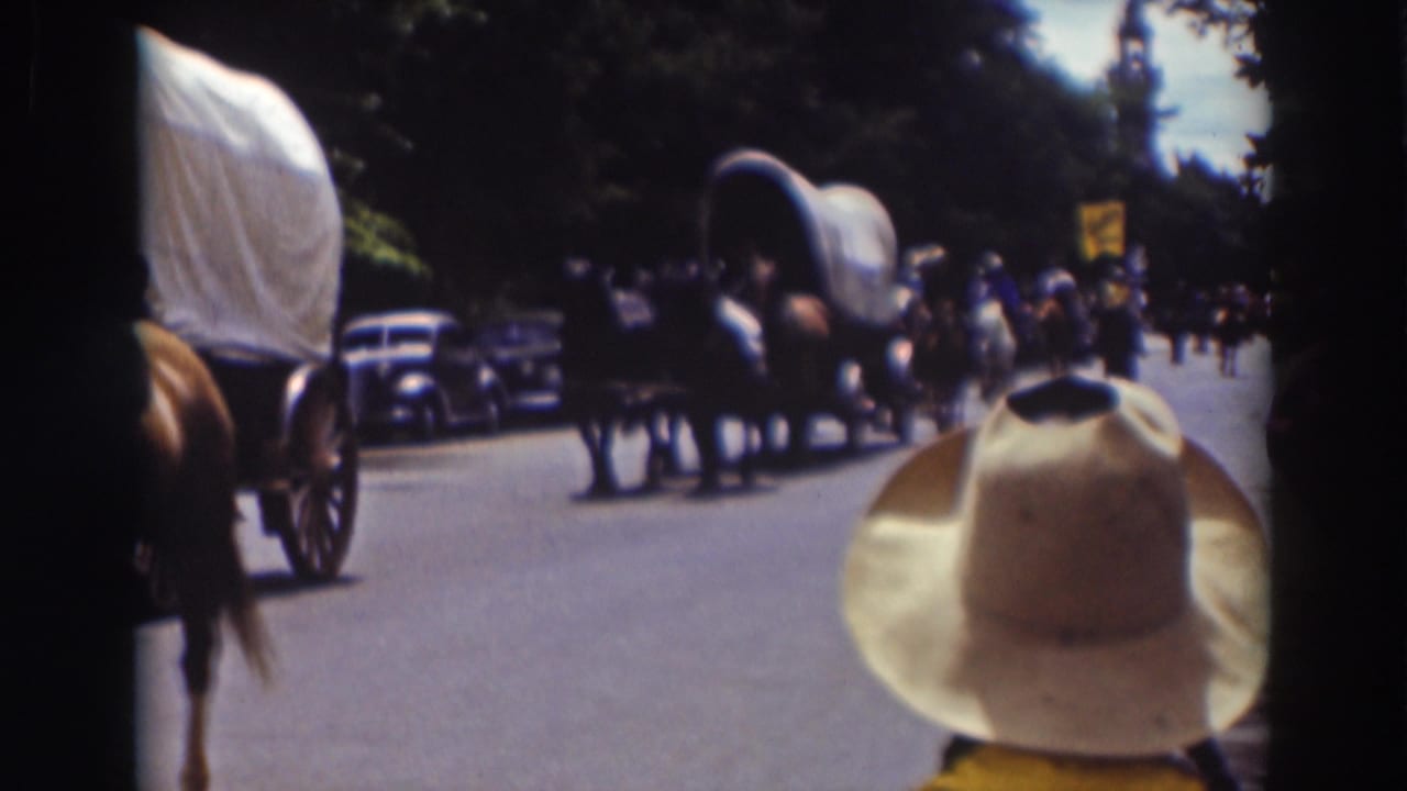 Late clip frame from the 1937 Truckee parade with covered wagons, horses, spectators, parked cars, and a cowboy hat in the foreground.