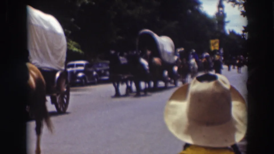 Late clip frame from the 1937 Truckee parade with covered wagons, horses, spectators, parked cars, and a cowboy hat in the foreground.