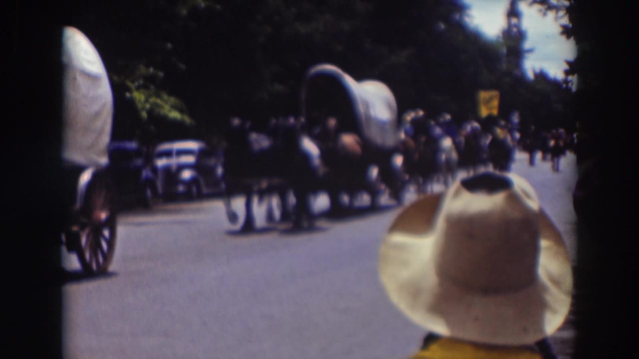 Covered wagon and horses pass spectators as a cowboy hat fills the foreground.