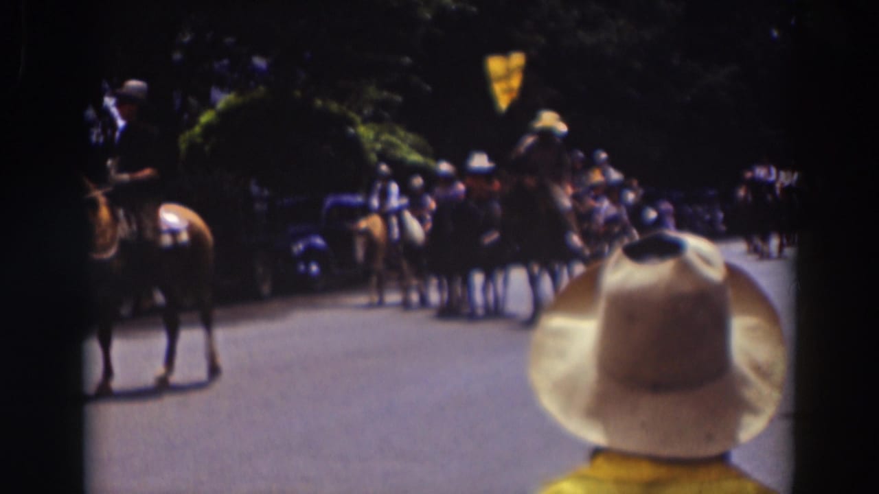 Spectator in a white cowboy hat watches horseback riders approach on a shaded street.