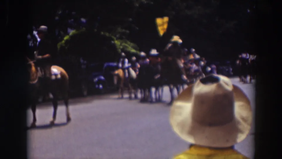 In 1937 Truckee, California, a spectator in a white cowboy hat stands in the foreground as riders move along a tree-lined parade route.