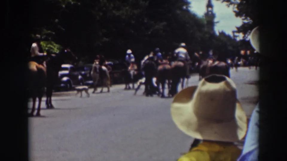 1937 Truckee 8mm frame showing riders on horseback gathered on a wooded road, viewed from behind a spectator in a light cowboy hat.
