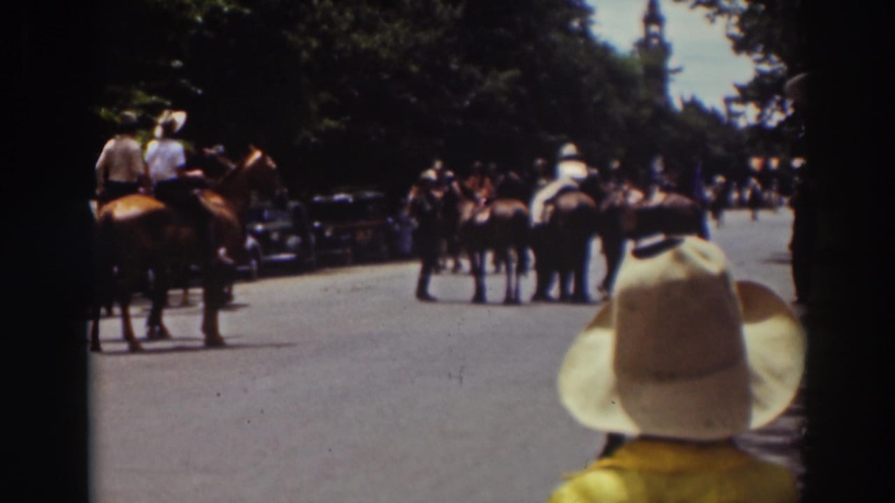 A child in a wide-brimmed hat watches mounted riders proceed down the street.
