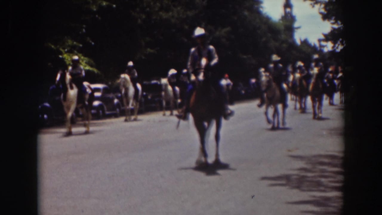 A mounted parade rider approaches in a 1937 Truckee street scene, with parked cars, spectators, and other riders behind.
