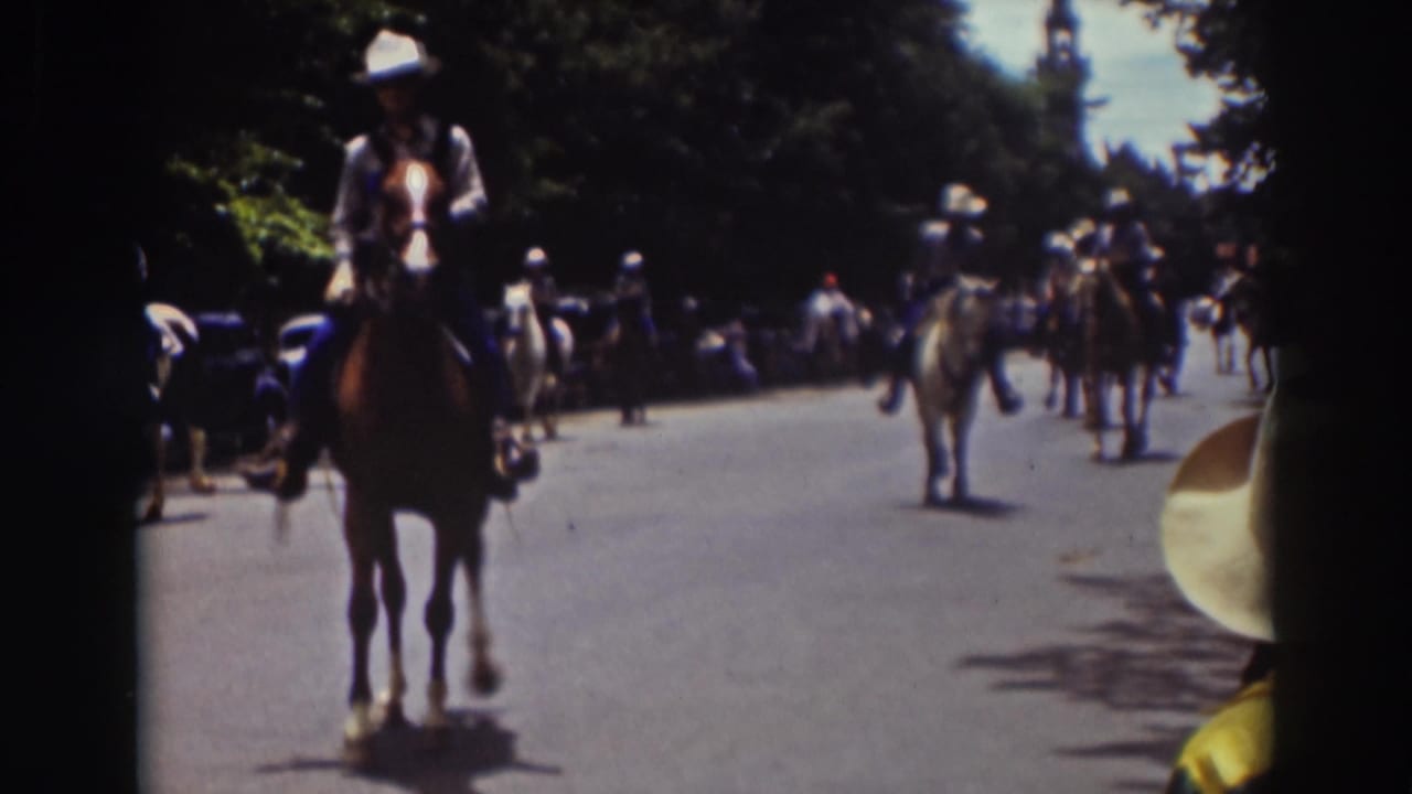 Mounted riders approach on a wide street with spectators and parked cars along the curb.