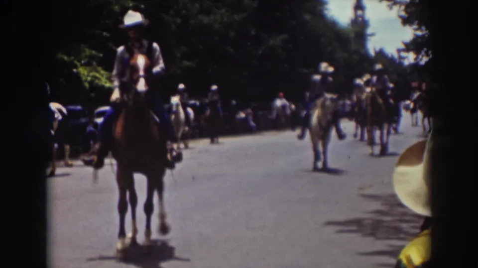 1937 Truckee home-movie frame showing a mounted parade moving down a broad tree-lined street with parked period cars.
