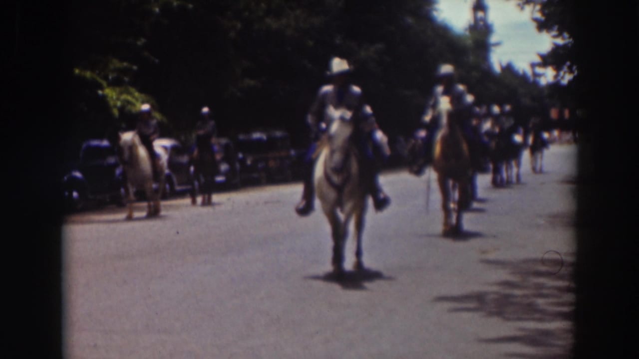 Mounted riders in white shirts approach on a tree-lined street with parked cars nearby.