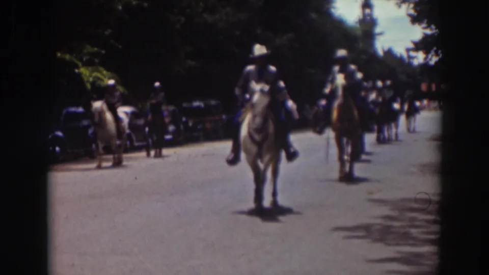 1937 Truckee parade footage shows horseback riders approaching the camera along a sunny street with early automobiles parked at the side.