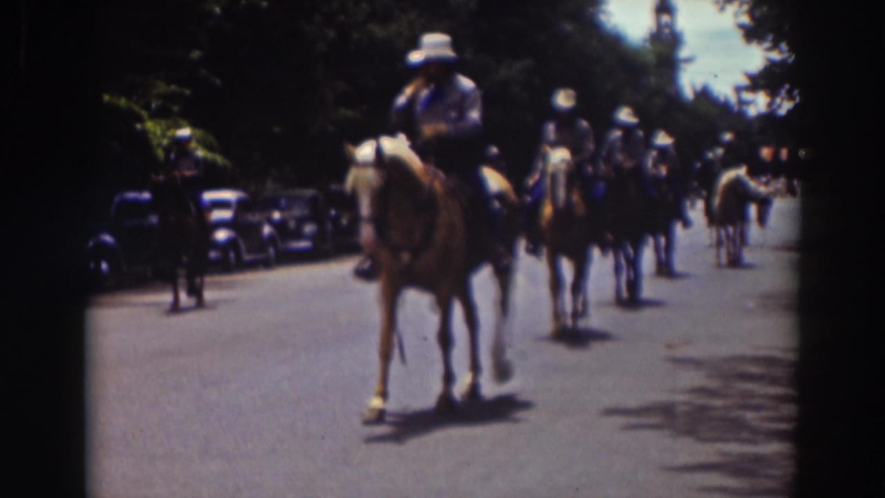 Mounted parade riders in white hats move along a sunny street lined with parked cars.