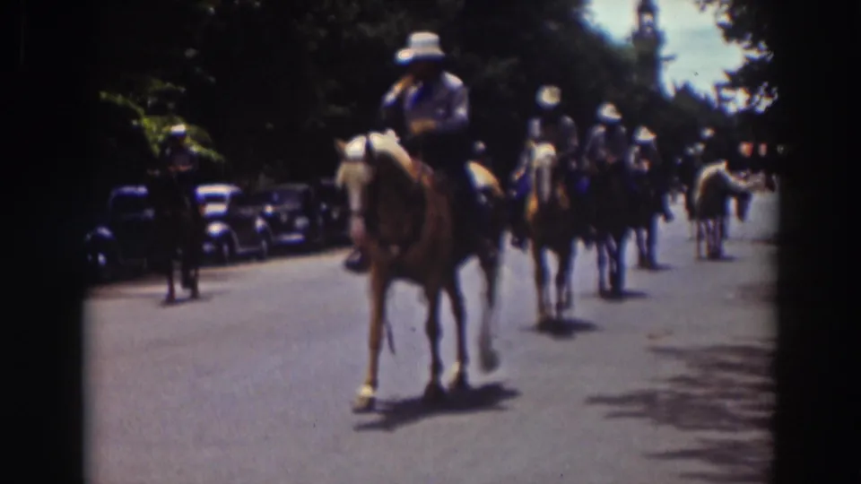 1937 Truckee home-movie frame showing horseback riders in white hats passing down a tree-lined street with parked cars nearby.