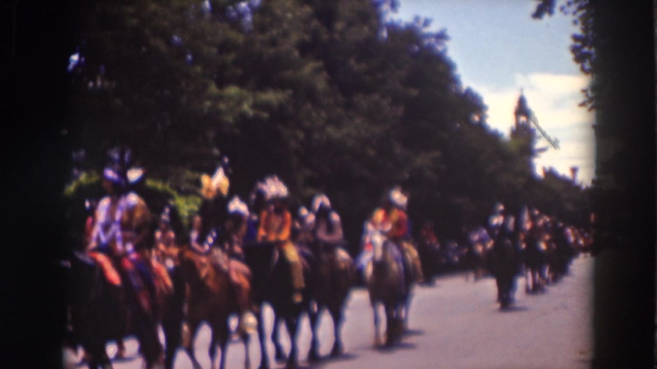 Horseback riders in feathered headdresses move down a tree-lined street.