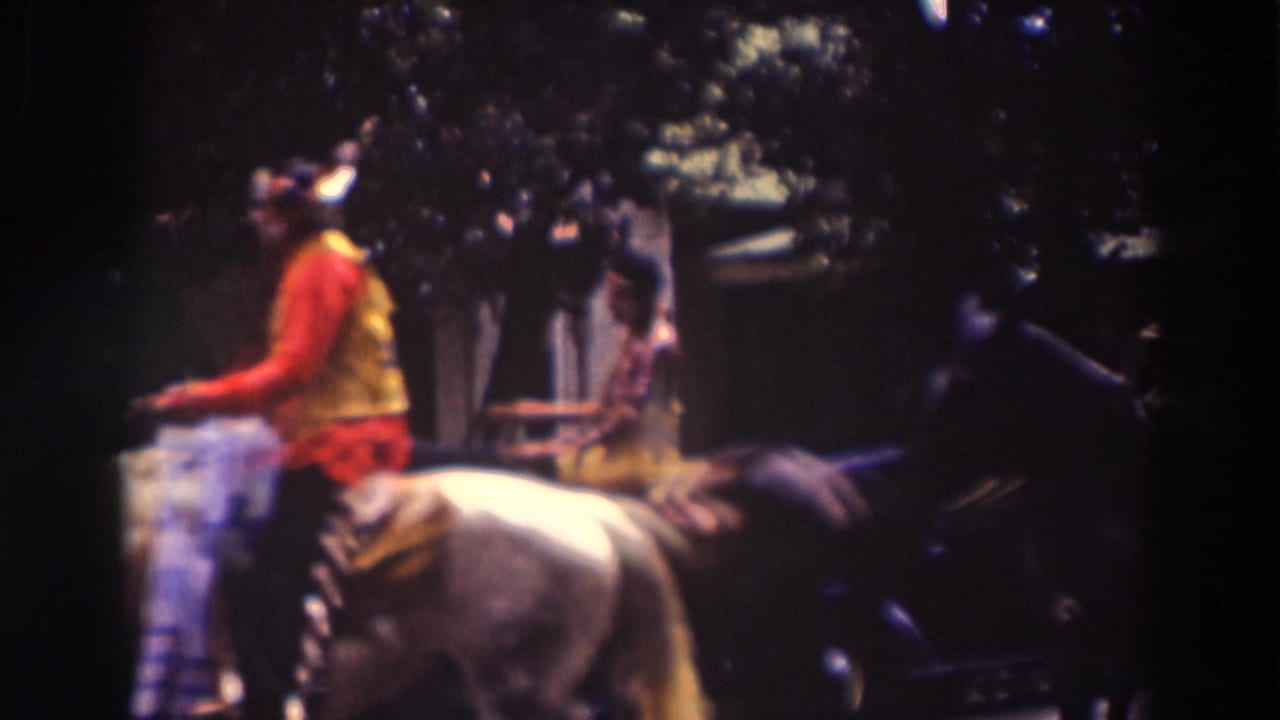 A parade rider in red and yellow clothing passes close by on a light-colored horse, with shaded spectators and buildings behind.