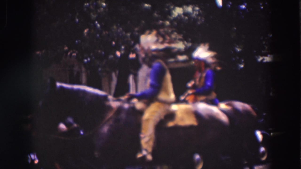 A rider in blue sleeves, yellow vest, and a feathered headdress passes on a dark horse, with parked period cars in the shade.