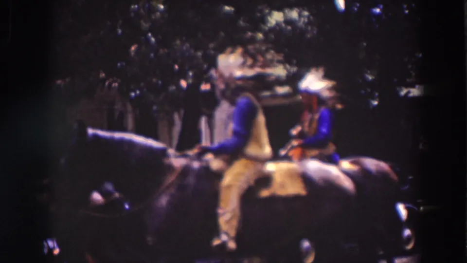 A rider in blue sleeves, yellow vest, and a feathered headdress passes on a dark horse, with parked period cars in the shade.