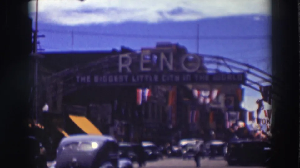 A Reno arch sign reading The Biggest Little City in the World spans a busy street with period cars and mountains behind it.