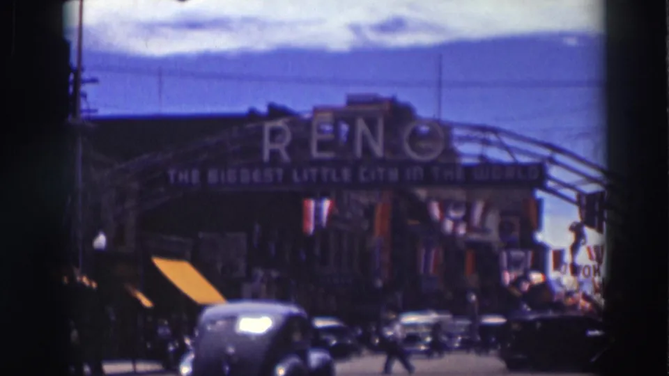 1937 home-movie frame showing cars and pedestrians on a commercial street beneath the Reno arch sign.