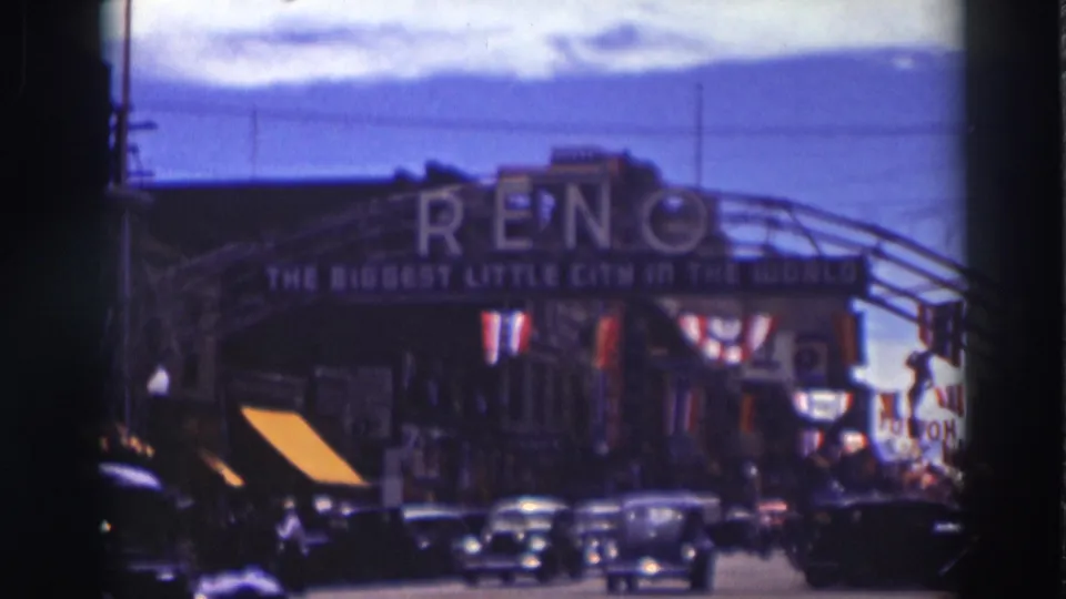 1937 home-movie frame showing multiple cars, pedestrians, and storefronts beneath the Reno arch sign in soft color 8mm.