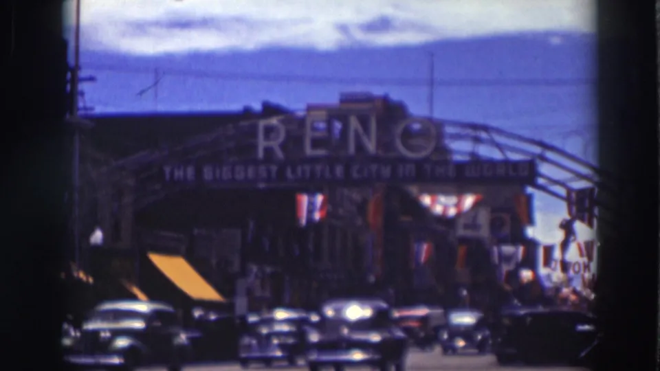 Soft 8mm frame of the Reno arch over a street lined with 1930s cars, flags, banners, and pedestrians along the sidewalk.