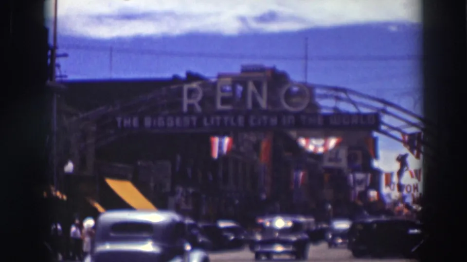 A car moves toward the Reno arch in a 1937 home-movie street view, with traffic, pedestrians, bunting, and flags visible around the gateway.