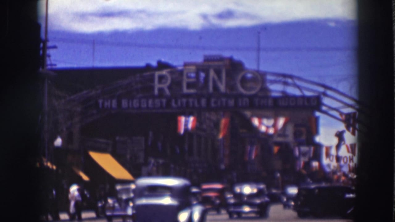 1937 travel footage view of cars moving under the Reno arch sign, with storefront awnings, flags, and pedestrians visible along the street.