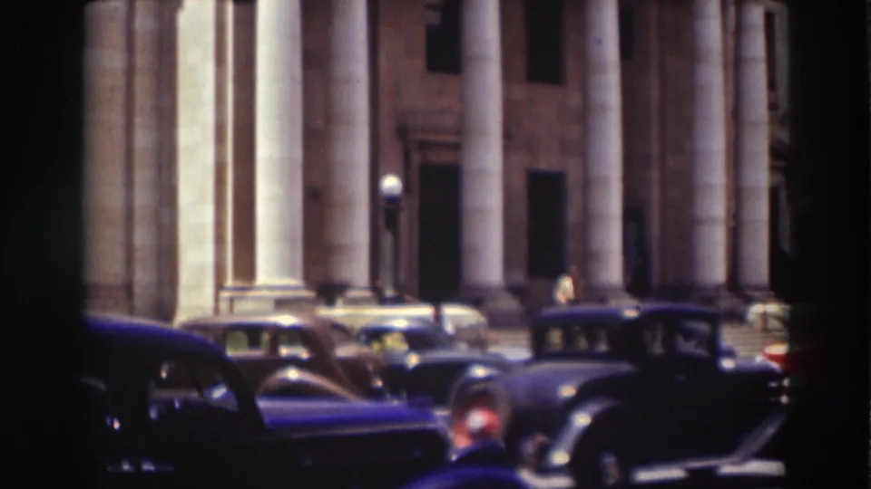 Cars and a few pedestrians appear near the entrance of a large classical building with tall columns and stone walls.