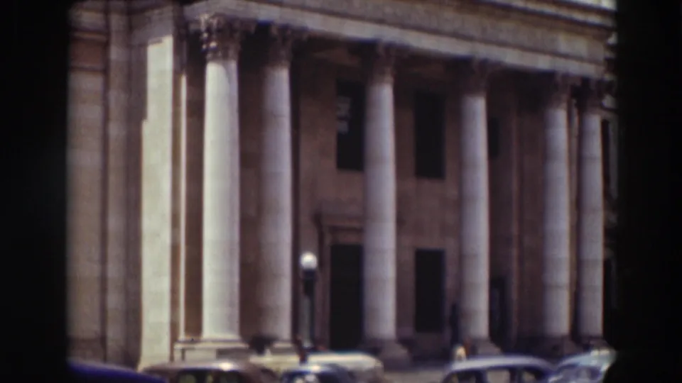 A handheld 8mm view frames the columns and entrance of a large stone building, with period cars along the curb in the foreground.