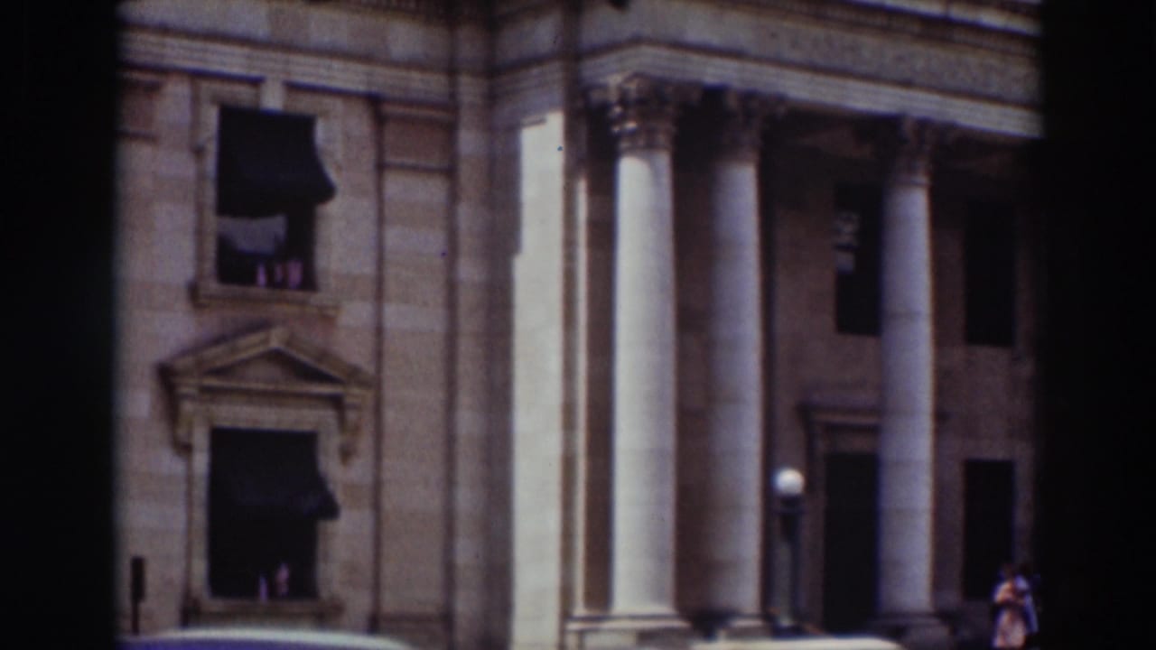 1937 Truckee frame showing a domed classical building with tall columns, stonework, dark awnings, and blue sky.