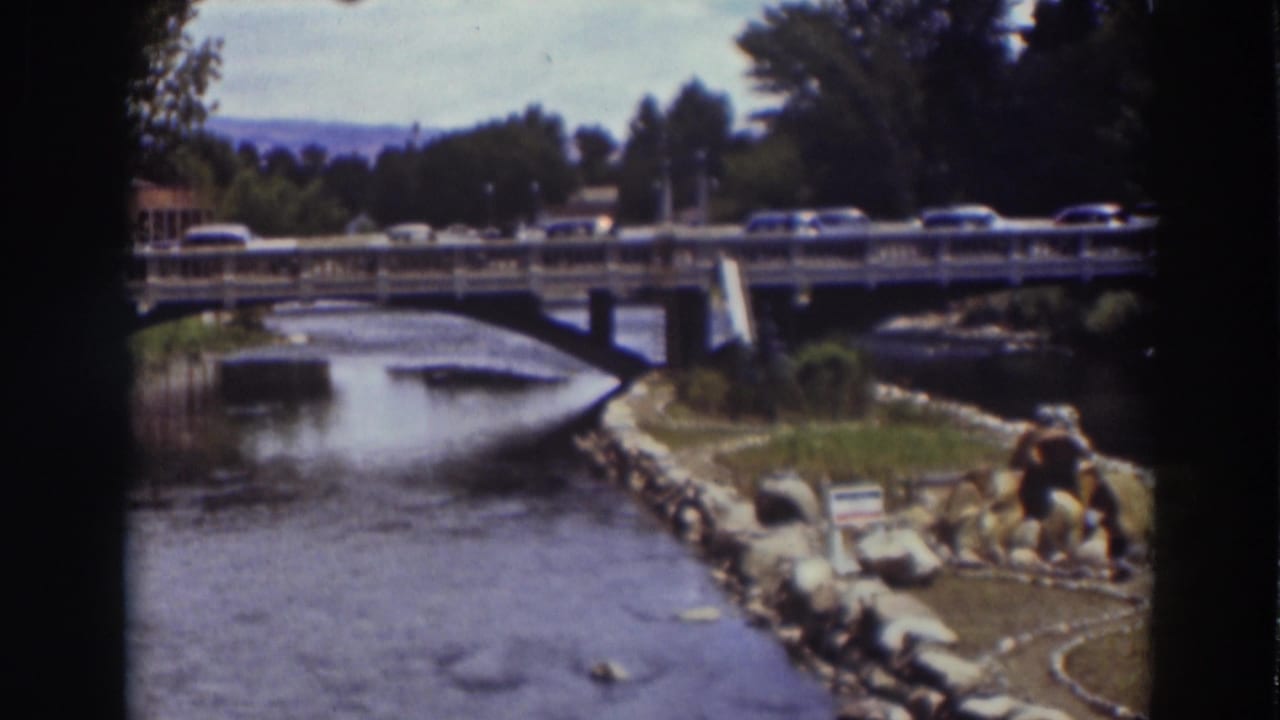 1937 Truckee frame with cars crossing the bridge, a stream below, and small people visible near the landscaped bank.