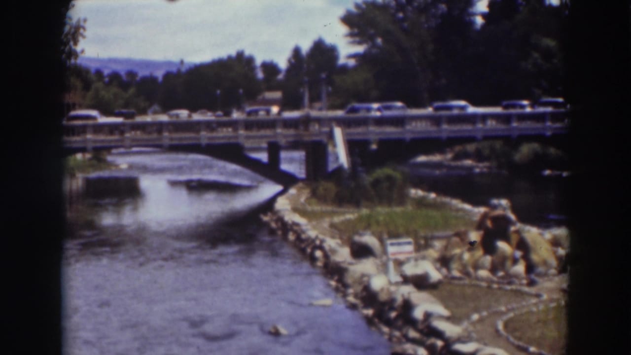 Cars cross a bridge over a river with rocks, grass, trees, and distant hills.