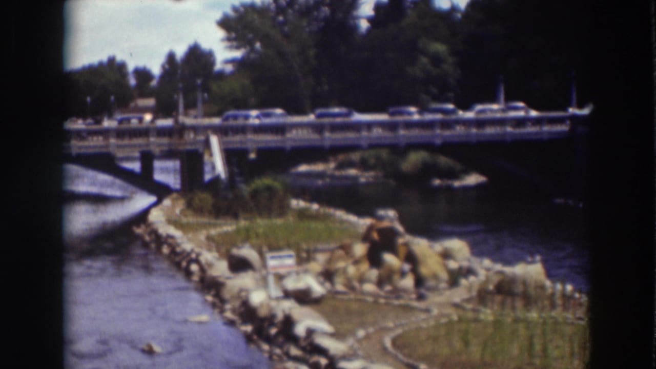 Home-movie frame from Truckee showing cars on a bridge, a river bend, landscaped rocks, grass, and dense trees.