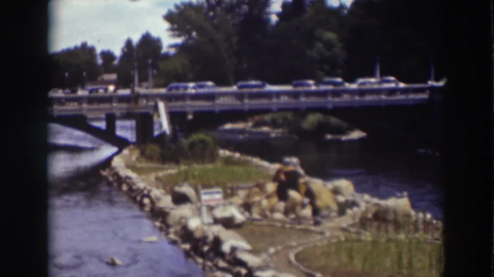 Home-movie frame from Truckee showing cars on a bridge, a river bend, landscaped rocks, grass, and dense trees.