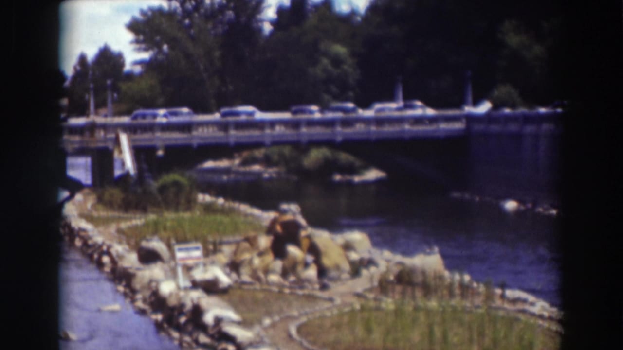 Late frame of the 1937 Truckee bridge scene, showing cars over the river and a landscaped bank in bright sun.
